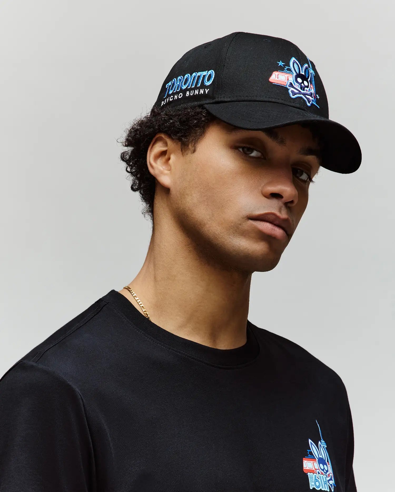 A young man wears the Psycho Bunny MENS TORONTO BASEBALL CAP (MO1500049) with an embroidered Bunny logo and adjustable strap, paired with a black tee featuring a blue and red chest graphic, posing against a plain light background.