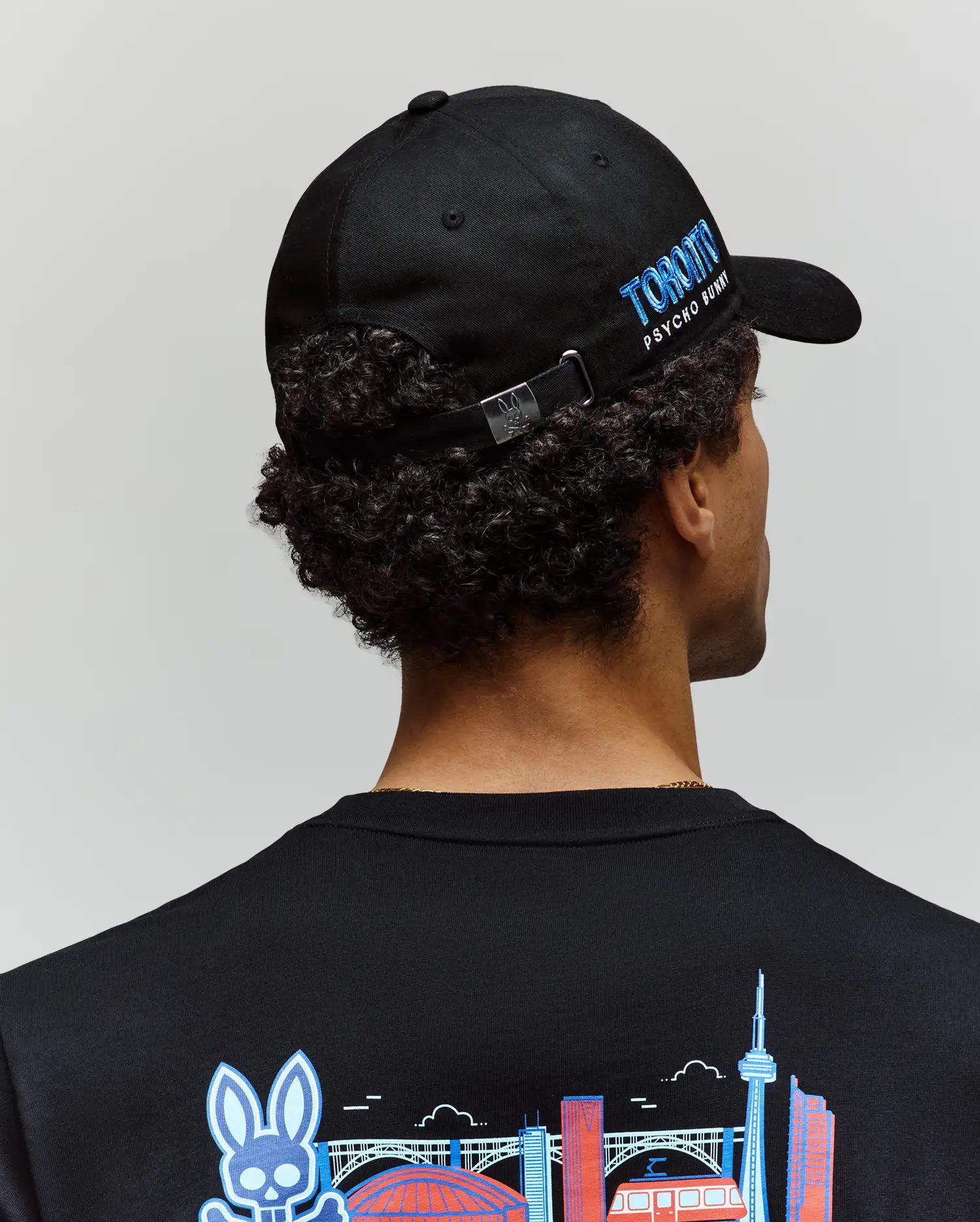 A person with curly hair, seen from behind, wears the Psycho Bunny MENS TORONTO BASEBALL CAP (MO1500049) featuring blue "TORONTO PSYCHO BUNNY" text and an embroidered shirt with colorful Toronto landmarks.