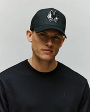 A man wearing the Psycho Bunny MENS AUGUSTE TRUCKER CAP (MO1500050), featuring a black design with a cartoon bunny chenille patch, and a black sweatshirt stands against a plain light background, looking directly at the camera.