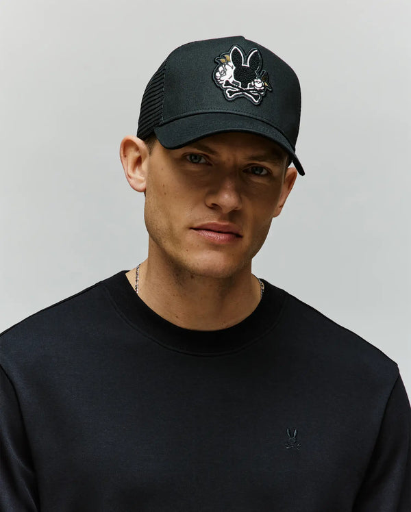 A man wearing the Psycho Bunny MENS AUGUSTE TRUCKER CAP (MO1500050), featuring a black design with a cartoon bunny chenille patch, and a black sweatshirt stands against a plain light background, looking directly at the camera.