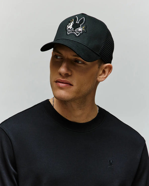 A man in a black sweatshirt and a Psycho Bunny MENS AUGUSTE TRUCKER CAP (MO1500050) with a cartoon bunny skull-and-crossbones chenille patch looks to the side against a plain light background.