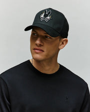 A man in a black sweatshirt and a Psycho Bunny MENS AUGUSTE TRUCKER CAP (MO1500050) with a cartoon bunny skull-and-crossbones chenille patch looks to the side against a plain light background.