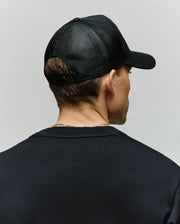 A person is shown from behind against a plain light background, wearing a black Psycho Bunny MENS AUGUSTE TRUCKER CAP (MO1500050) with an adjustable snapback and mesh design, paired with a black shirt.
