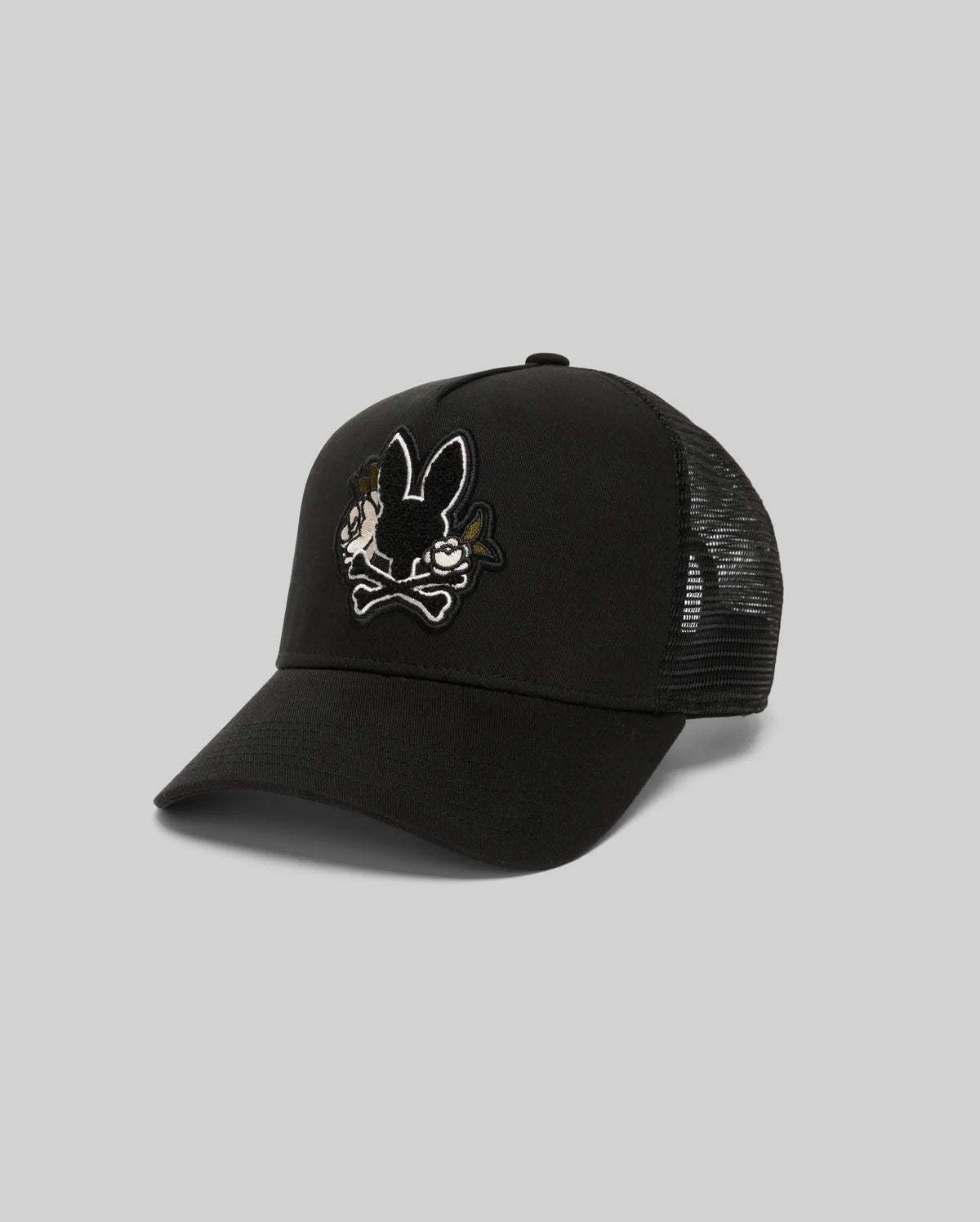 The Psycho Bunny MENS AUGUSTE TRUCKER CAP (MO1500050) is a black mesh-back hat with an embroidered cartoon bunny and crossbones chenille patch on the front. Features an adjustable snapback for comfort. Shown on a light gray background.