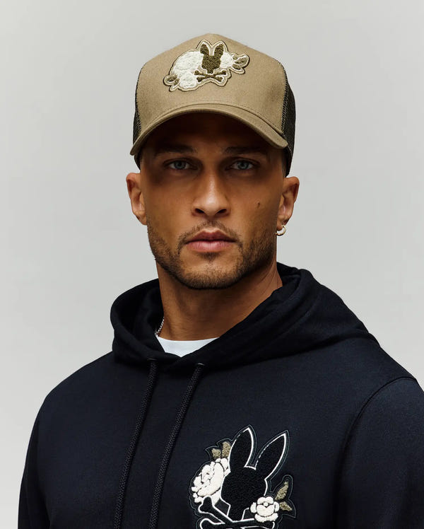 A man wears the Psycho Bunny MENS AUGUSTE TRUCKER CAP (MO1500050), a tan hat with a chenille bunny patch, and a black hoodie with matching bunny and flower designs, standing against a simple light background.