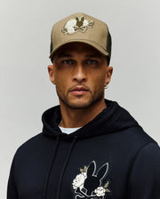 A man wears the Psycho Bunny MENS AUGUSTE TRUCKER CAP (MO1500050), a tan hat with a chenille bunny patch, and a black hoodie with matching bunny and flower designs, standing against a simple light background.