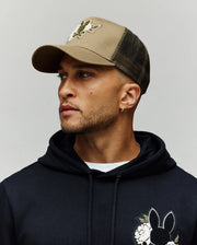 A man with a short beard and gold hoop earring wears the Psycho Bunny MENS AUGUSTE TRUCKER CAP (MO1500050), beige and black with a mesh back and snapback closure, along with a black hoodie featuring a floral bunny chenille patch.