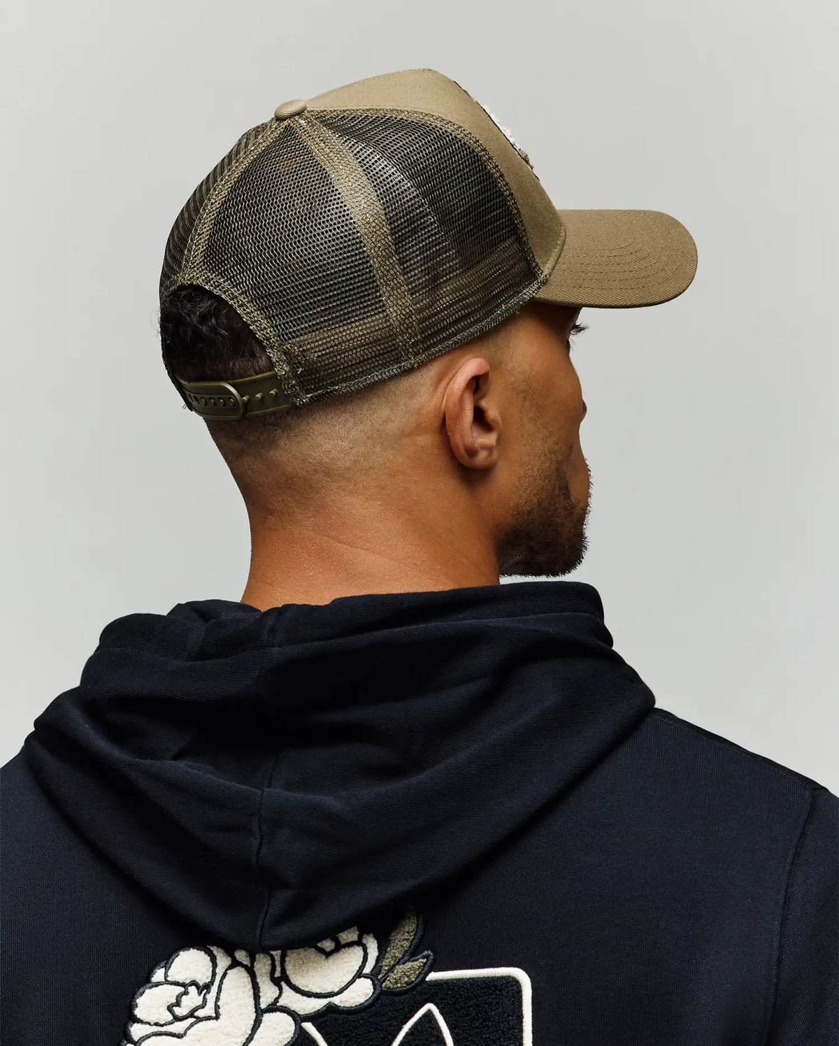 A man, shown from behind against a plain gray background, wears a black hoodie with a white floral back design and the Psycho Bunny MENS AUGUSTE TRUCKER CAP - MO1500050 in olive green mesh with snapback closure.