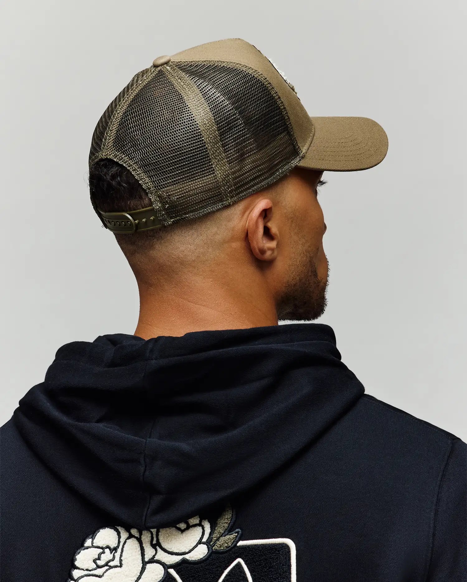 A man, shown from behind against a plain gray background, wears a black hoodie with a white floral back design and the Psycho Bunny MENS AUGUSTE TRUCKER CAP - MO1500050 in olive green mesh with snapback closure.