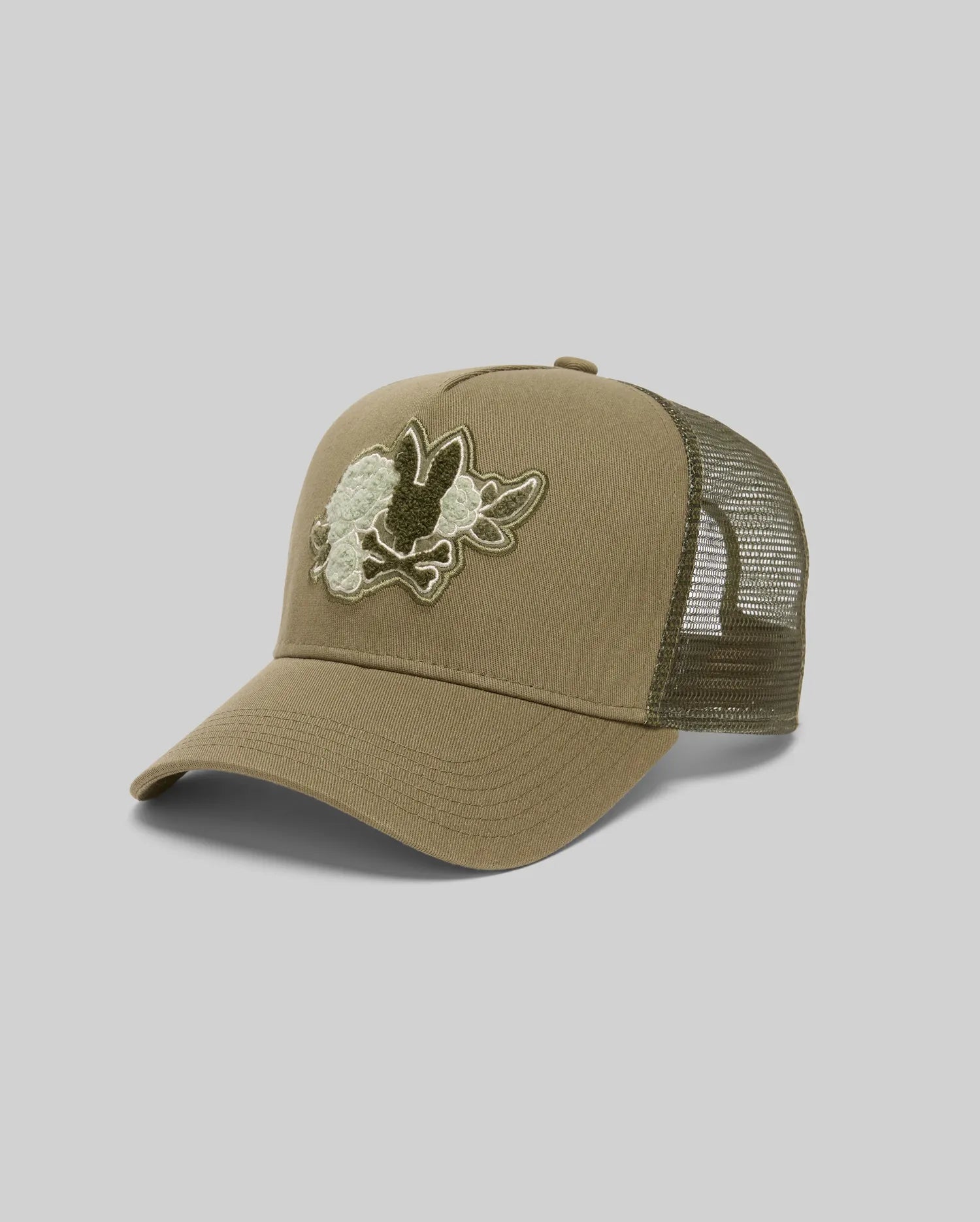 The Psycho Bunny MENS AUGUSTE TRUCKER CAP (MO1500050) is a khaki green trucker hat with a mesh back, snapback closure, and an embroidered triceratops skull patch on the front panel, shown on a light gray background.