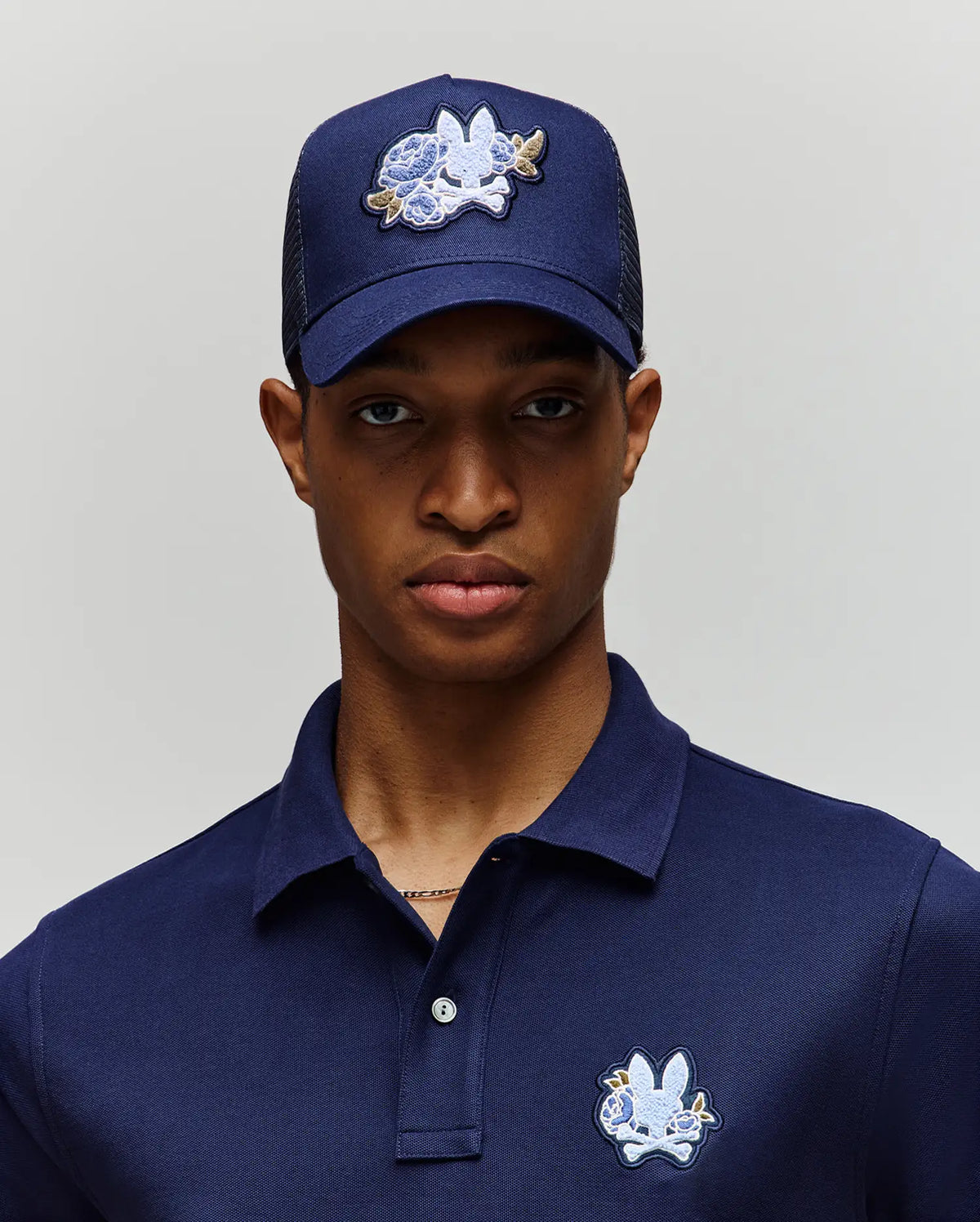 A person wears the Psycho Bunny MENS AUGUSTE TRUCKER CAP - MO1500050 in navy blue, featuring a white and blue chenille patchwork bunny design and an adjustable snapback for comfort, paired with a matching polo shirt against a plain background.