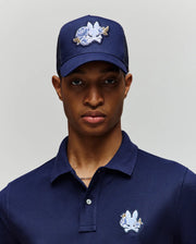 A person wears the Psycho Bunny MENS AUGUSTE TRUCKER CAP - MO1500050 in navy blue, featuring a white and blue chenille patchwork bunny design and an adjustable snapback for comfort, paired with a matching polo shirt against a plain background.