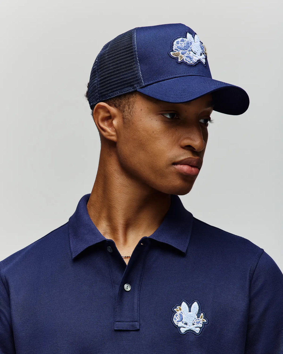 A young man wears a navy blue polo shirt and the Psycho Bunny MENS AUGUSTE TRUCKER CAP (MO1500050), both featuring an embroidered white and blue cartoon bunny logo, as he looks slightly left against a plain light background.