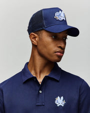 A young man wears a navy blue polo shirt and the Psycho Bunny MENS AUGUSTE TRUCKER CAP (MO1500050), both featuring an embroidered white and blue cartoon bunny logo, as he looks slightly left against a plain light background.