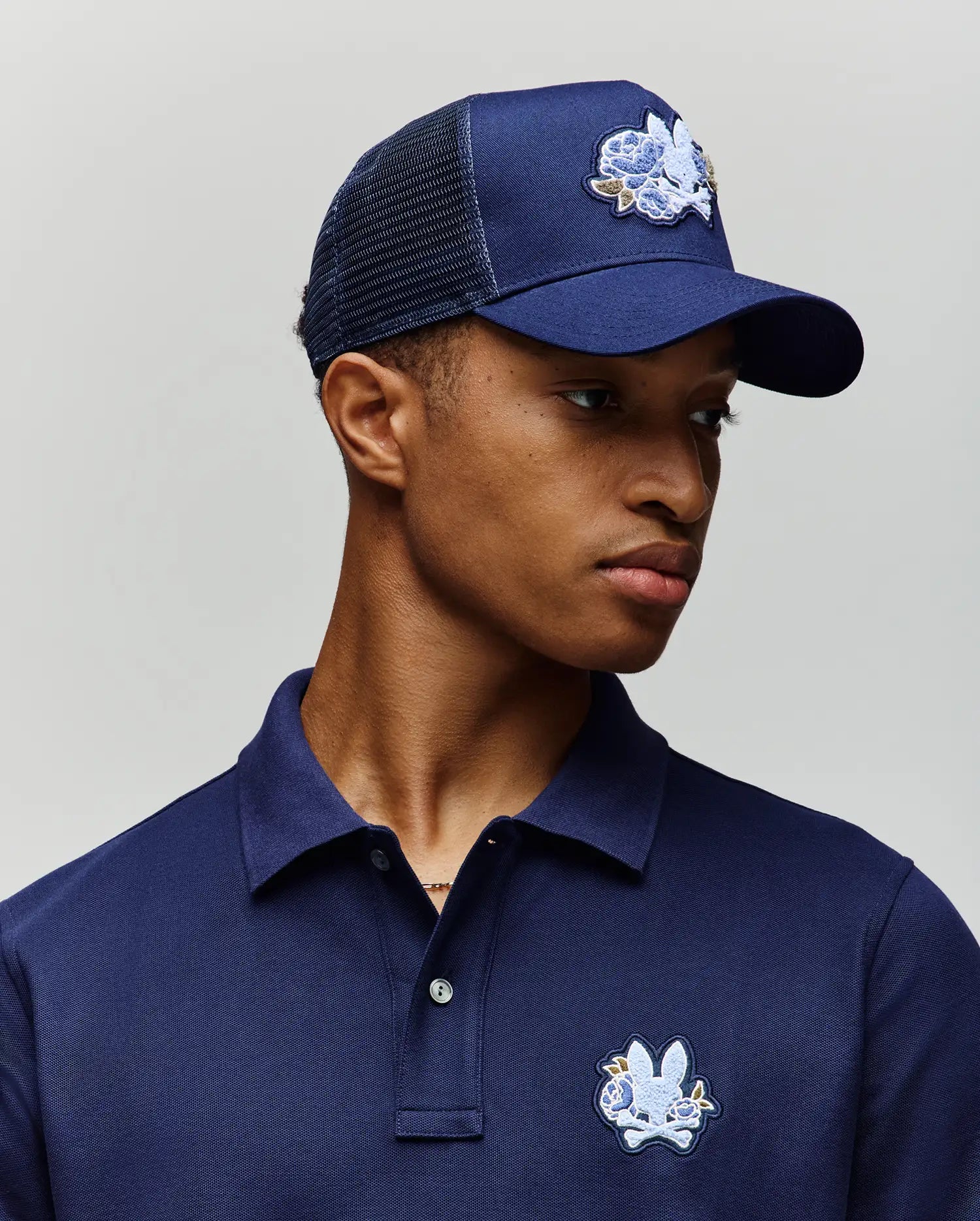 A young man wears a navy blue polo shirt and the Psycho Bunny MENS AUGUSTE TRUCKER CAP (MO1500050), both featuring an embroidered white and blue cartoon bunny logo, as he looks slightly left against a plain light background.