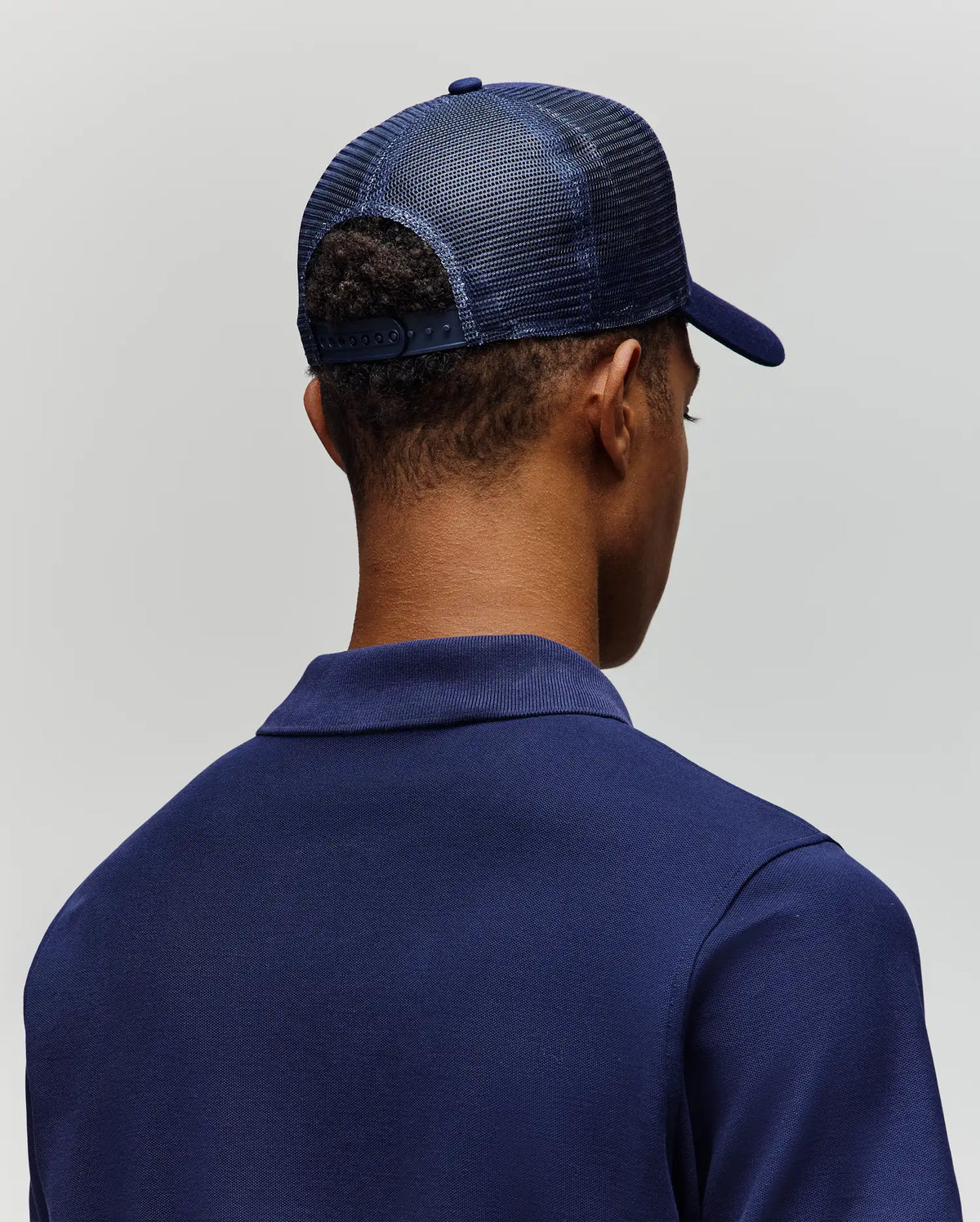 A person is shown from behind against a light gray background, wearing a Psycho Bunny MENS AUGUSTE TRUCKER CAP - MO1500050, a dark blue mesh trucker hat with an adjustable snapback, and a blue collared shirt.