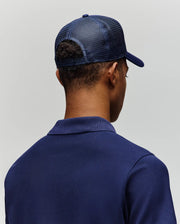 A person is shown from behind against a light gray background, wearing a Psycho Bunny MENS AUGUSTE TRUCKER CAP - MO1500050, a dark blue mesh trucker hat with an adjustable snapback, and a blue collared shirt.