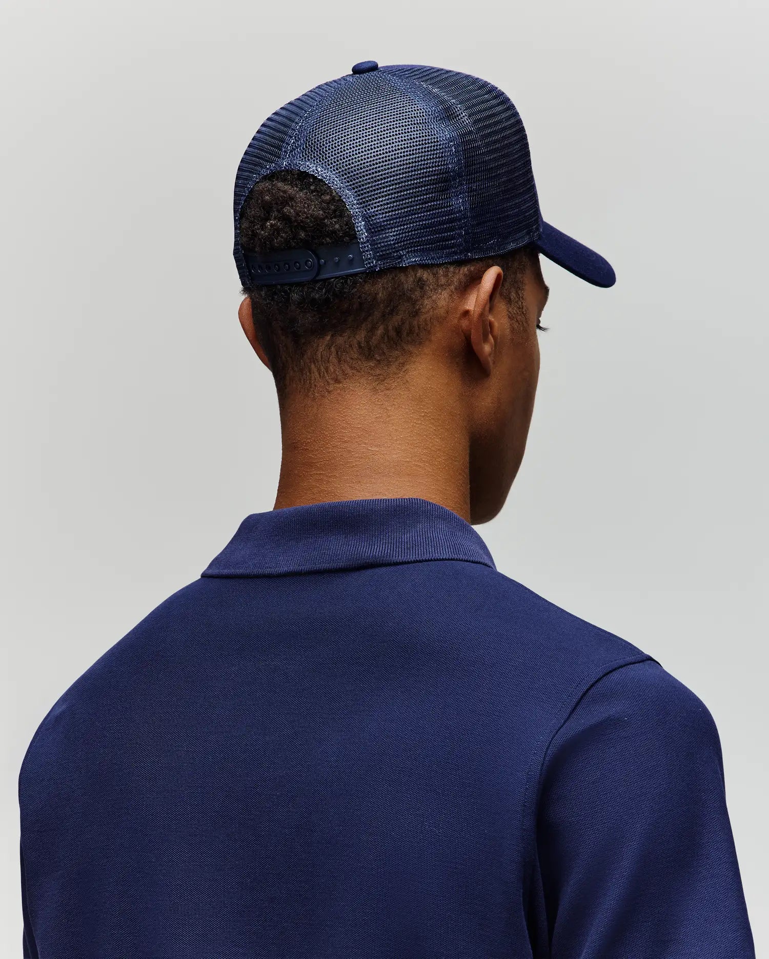 A person is shown from behind against a light gray background, wearing a Psycho Bunny MENS AUGUSTE TRUCKER CAP - MO1500050, a dark blue mesh trucker hat with an adjustable snapback, and a blue collared shirt.
