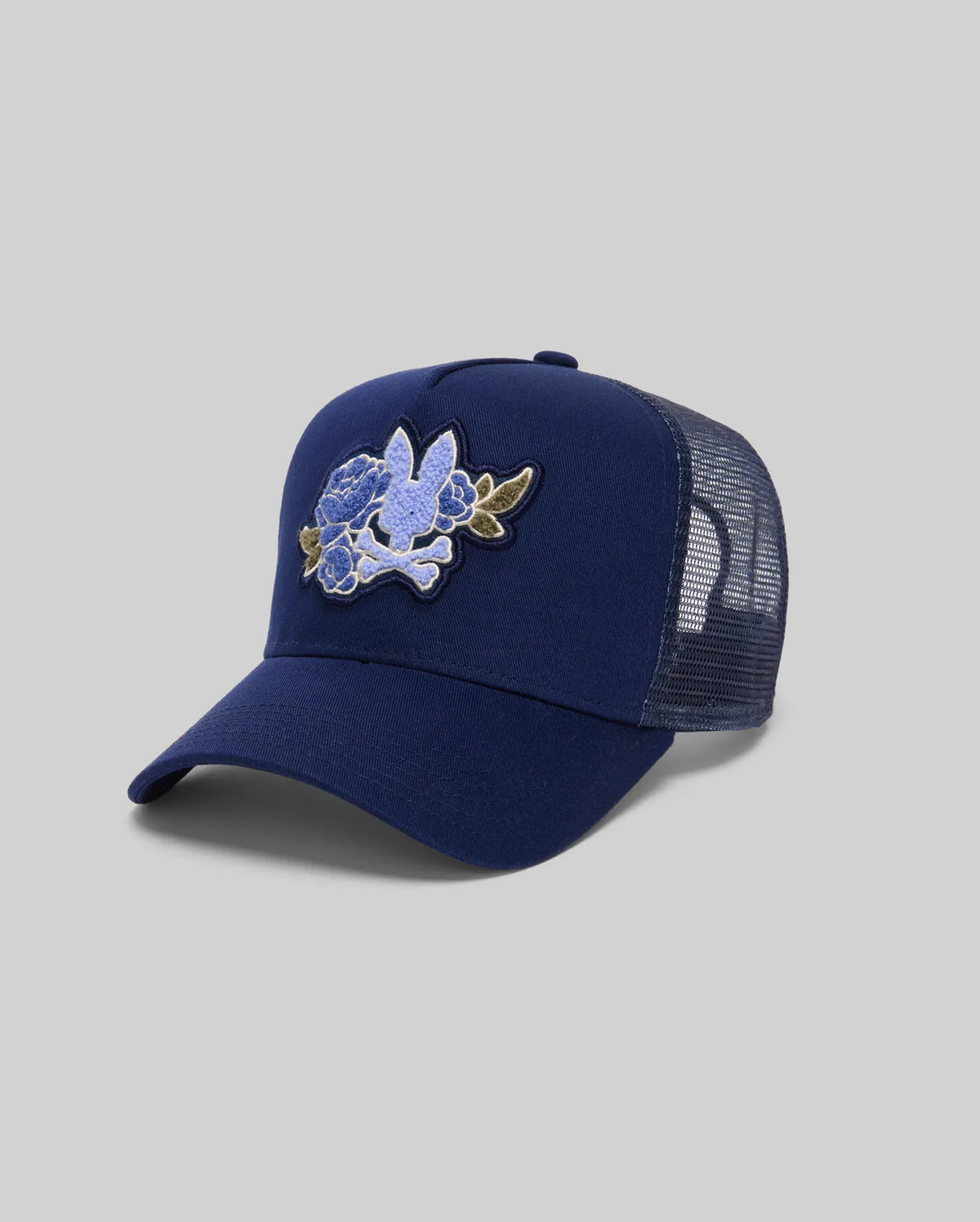 The Psycho Bunny MENS AUGUSTE TRUCKER CAP (MO1500050) is a navy blue hat with mesh back, adjustable snapback, and a chenille patchwork floral design of blue flowers and green leaves on the front, shown on a grey background.