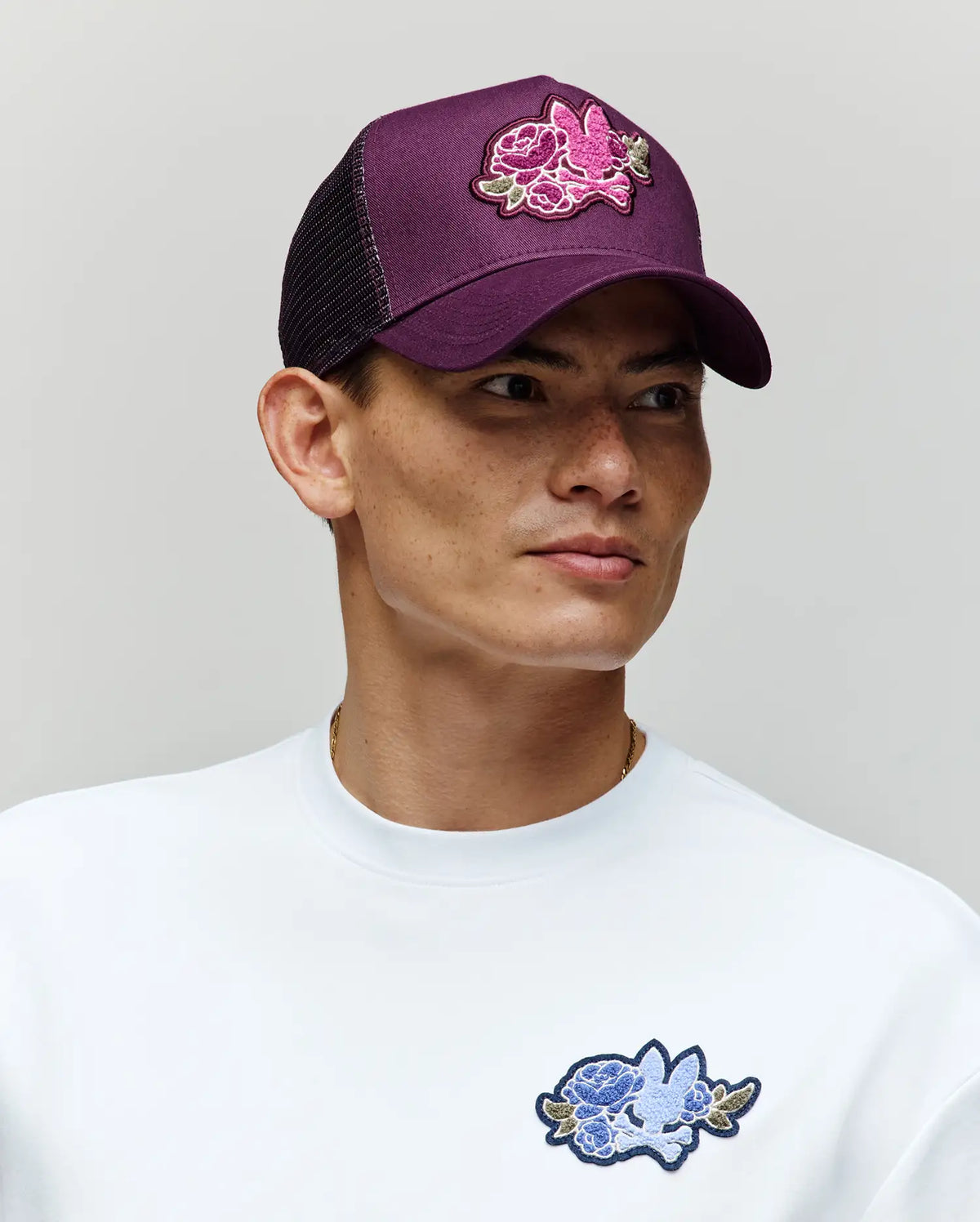 A person wears the Psycho Bunny MENS AUGUSTE TRUCKER CAP (MO1500050), a purple adjustable snapback with a pink chenille floral patch, paired with a white sweatshirt featuring a blue floral patch, looking to the side against a neutral background.