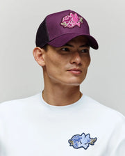 A person wears the Psycho Bunny MENS AUGUSTE TRUCKER CAP (MO1500050), a purple adjustable snapback with a pink chenille floral patch, paired with a white sweatshirt featuring a blue floral patch, looking to the side against a neutral background.