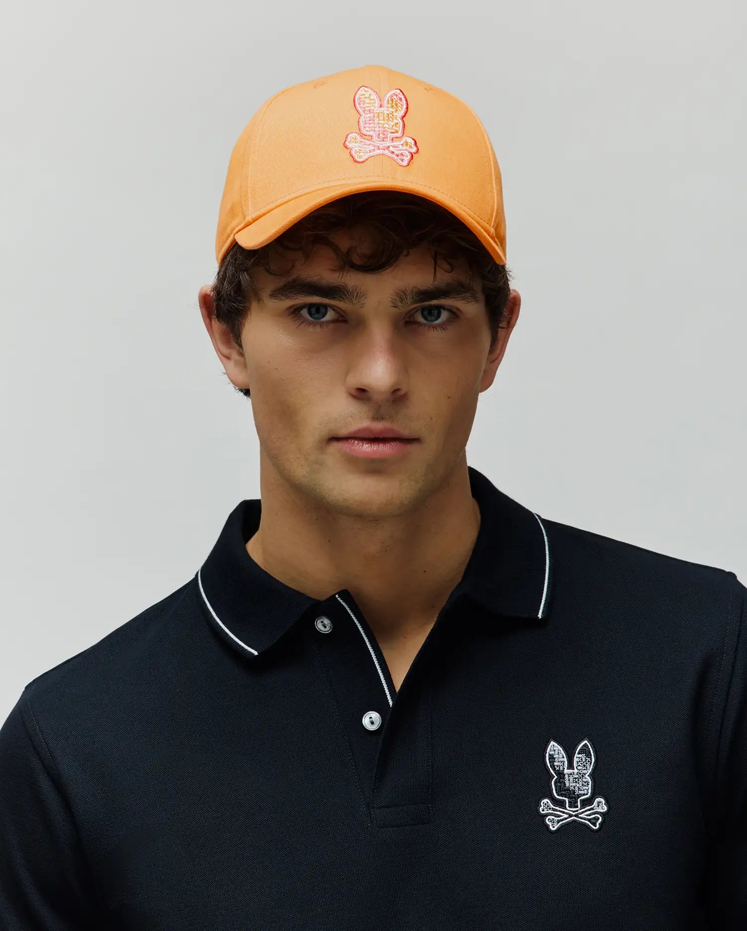Black pique polo shirt with white tipping and a textured Psycho Bunny skull logo on the chest, paired with an orange Mens Olivier Baseball Cap featuring a pink and white embroidered bunny skull.