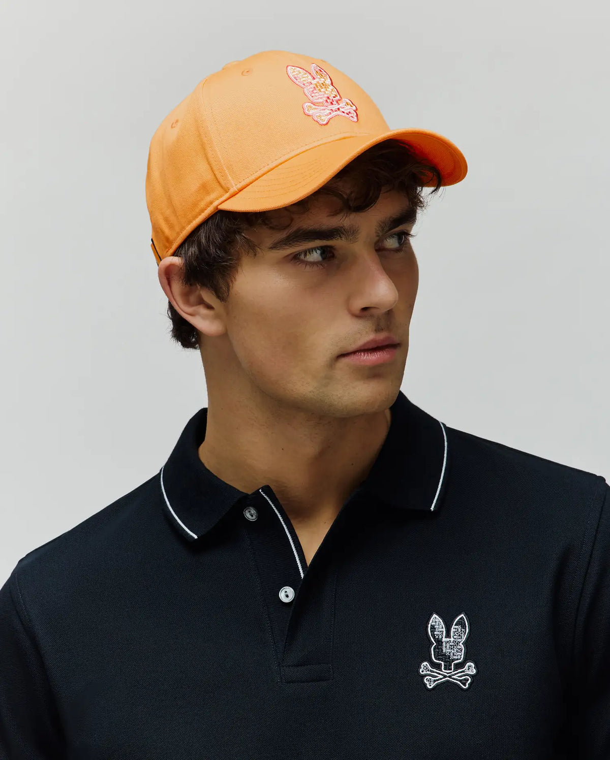 Black classic fit pique polo with white collar piping and a textured embroidered Psycho Bunny skull logo on chest. Orange Mens Olivier Baseball Cap with a multi-color embroidered bunny skull logo.
