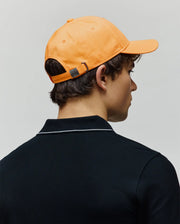 Mens Olivier Baseball Cap in bright orange cotton twill with a small debossed Psycho Bunny skull logo on the back buckle, paired with a dark navy pique polo shirt featuring a white striped collar.