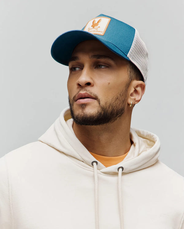 Mens Adamo Trucker Cap with a teal front panel, off-white mesh back, and a tan patch featuring the orange Psycho Bunny skull logo, paired with a relaxed fit cream french terry hoodie.