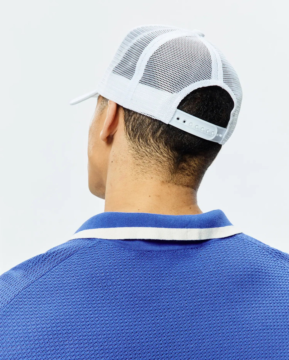 White Mens Adamo Trucker Cap with mesh back and snap closure, paired with a royal blue pique polo shirt featuring a blue and white striped collar. (3)
