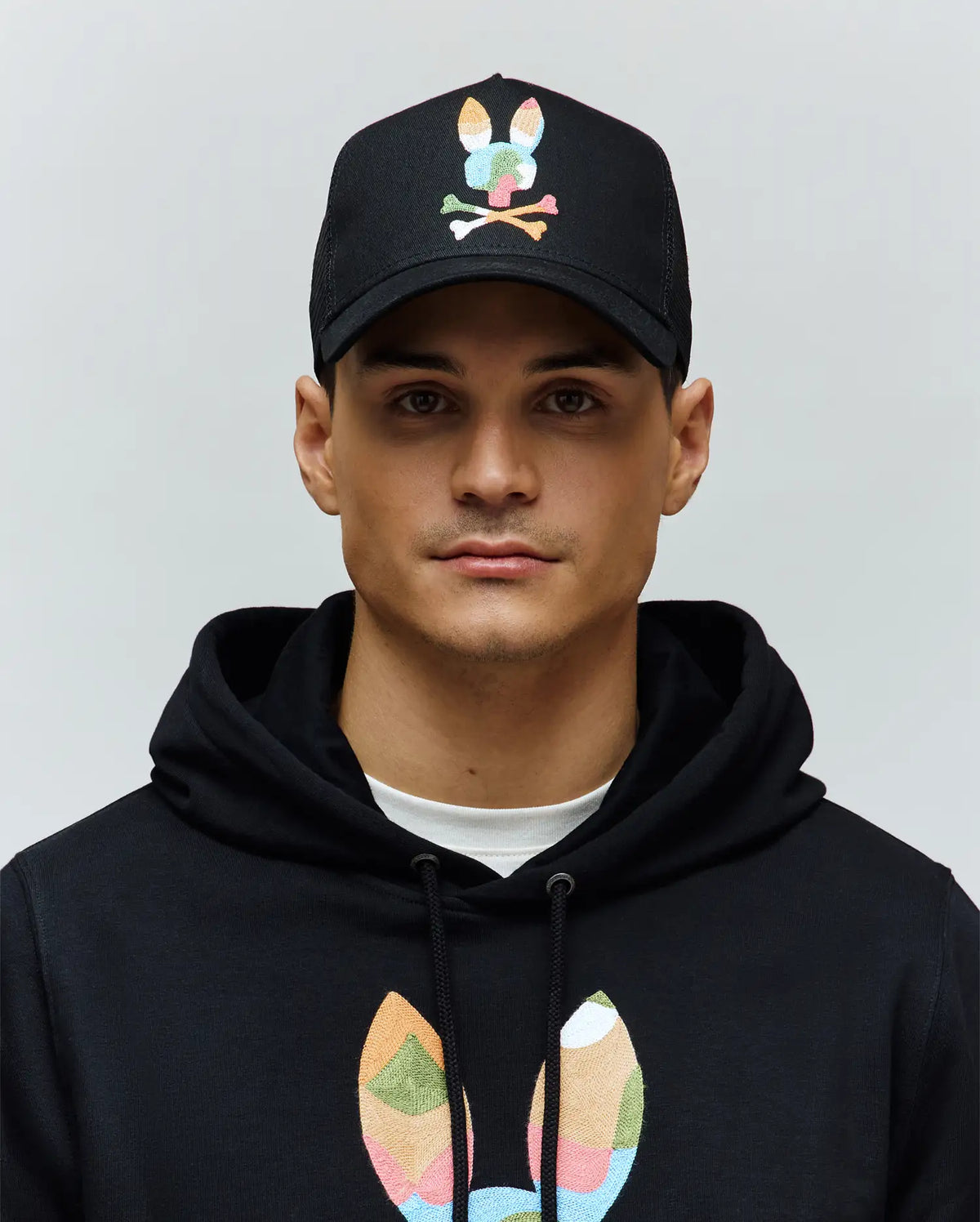 Black Mens Ara Trucker Cap with a vibrant, multi-color embroidered Psycho Bunny skull logo on the front, paired with a black hoodie featuring a large, matching colorful embroidered bunny skull on the chest.