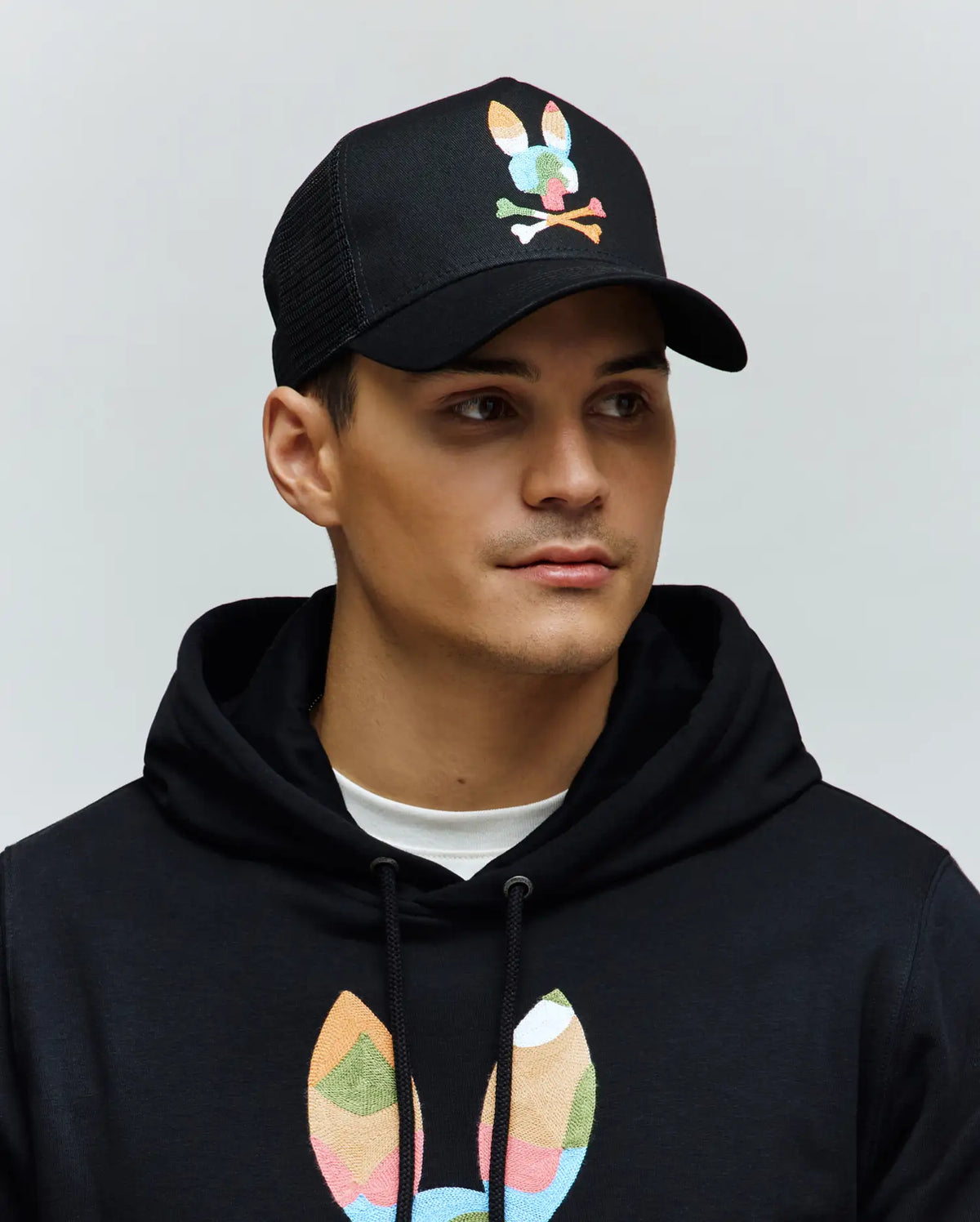 Mens Ara Trucker Cap in black with a large multicolor embroidered Psycho Bunny skull logo, paired with a black french terry hoodie featuring a matching large multicolor bunny skull graphic.