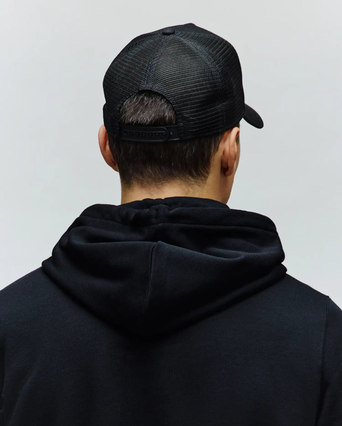 Black Mens Ara Trucker Cap featuring a breathable mesh back and adjustable snap closure, worn with a relaxed fit black Psycho Bunny hoodie crafted from soft jersey fabric.