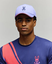 A person wears the Psycho Bunny MENS OFFICIAL US OPEN AVERY NET SPORT CAP—a light purple, breathable cap with a bunny and crossbones logo—and a blue athletic shirt with pink and red diagonal stripes and a matching bunny logo.
