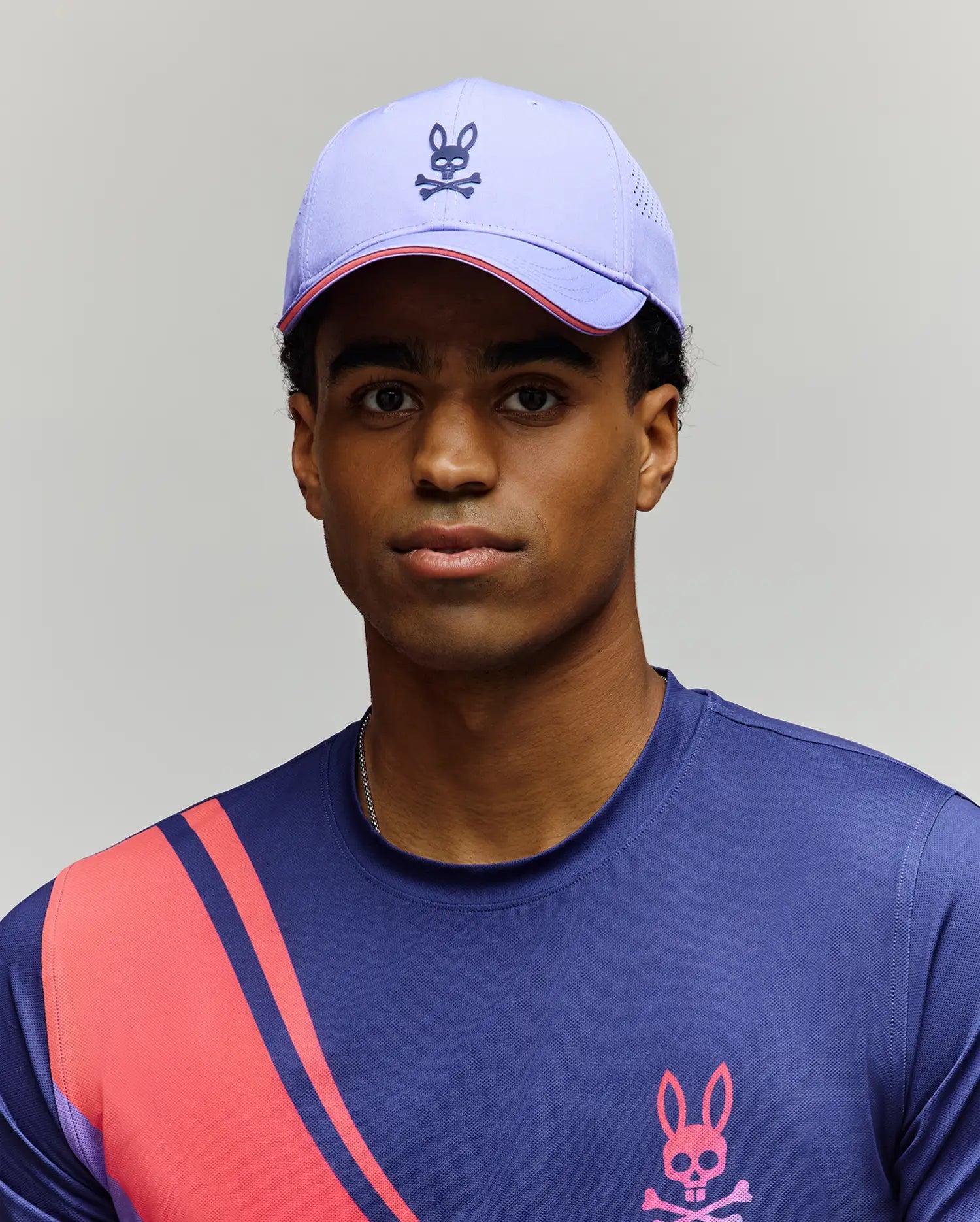A person wears the Psycho Bunny MENS OFFICIAL US OPEN AVERY NET SPORT CAP—a light purple, breathable cap with a bunny and crossbones logo—and a blue athletic shirt with pink and red diagonal stripes and a matching bunny logo.