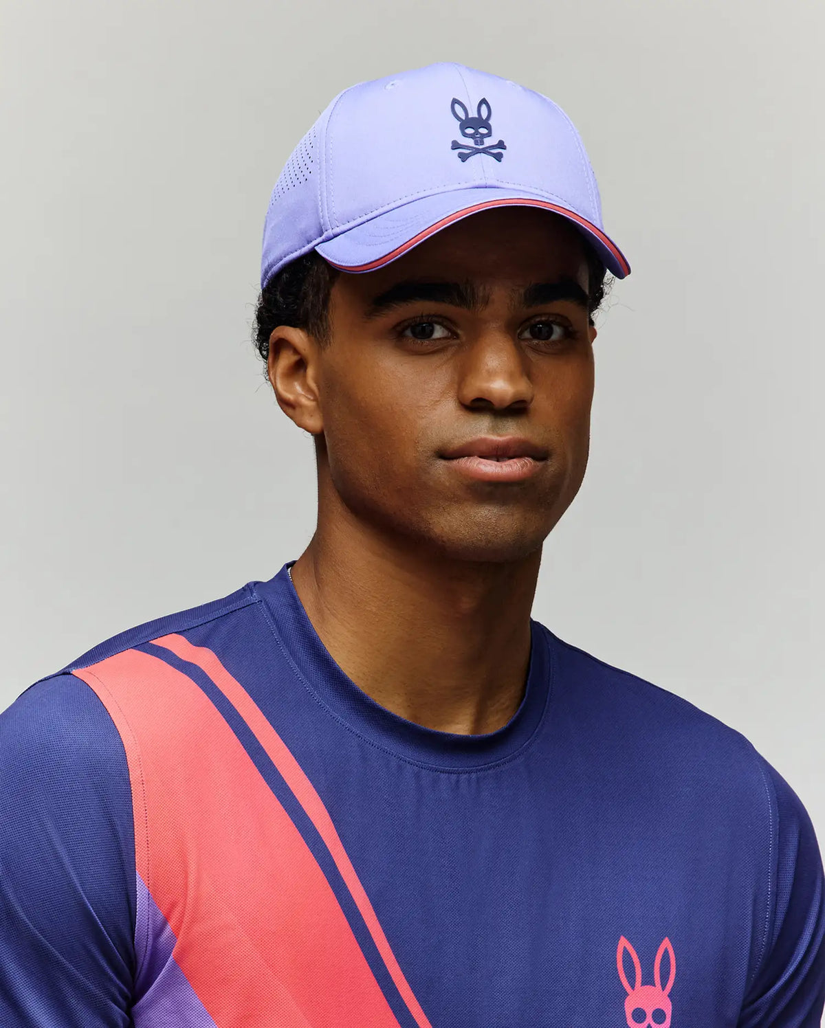 A man in a purple and pink Psycho Bunny MENS OFFICIAL US OPEN AVERY NET SPORT CAP (MO1500086), plus a blue shirt with pink and red diagonal stripes and a matching bunny logo, poses against a plain light background.
