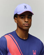 A man in a purple and pink Psycho Bunny MENS OFFICIAL US OPEN AVERY NET SPORT CAP (MO1500086), plus a blue shirt with pink and red diagonal stripes and a matching bunny logo, poses against a plain light background.