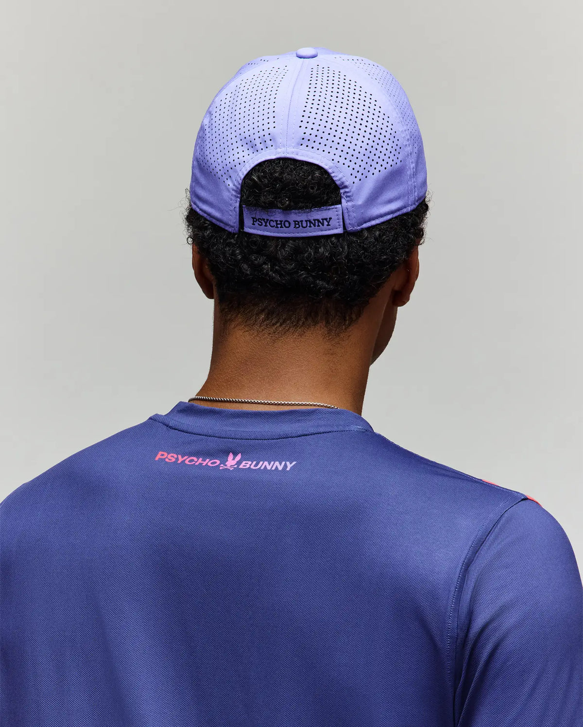 A man is seen from behind wearing a light purple MENS OFFICIAL US OPEN AVERY NET SPORT CAP - MO1500086 by Psycho Bunny and a blue Psycho Bunny shirt, set against a plain gray background to showcase the sporty cap’s stylish, breathable design.