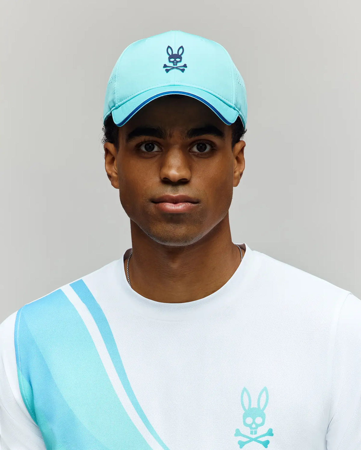 A person wears the MENS OFFICIAL US OPEN AVERY NET SPORT CAP by Psycho Bunny—a light blue, breathable hat with a bunny and crossbones logo—and a white shirt with blue stripes and matching bunny logo against a plain light gray background.