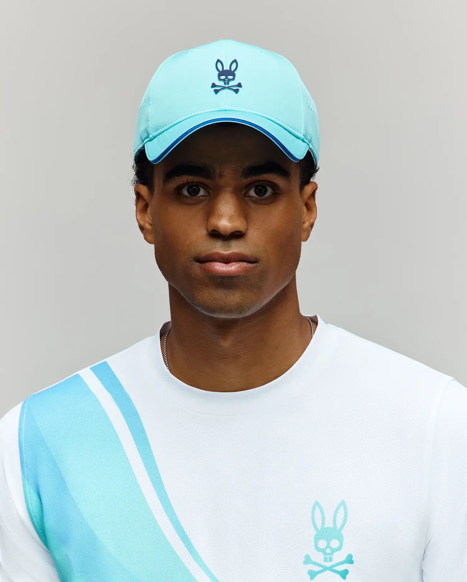 A person wears the MENS OFFICIAL US OPEN AVERY NET SPORT CAP by Psycho Bunny—a light blue, breathable hat with a bunny and crossbones logo—and a white shirt with blue stripes and matching bunny logo against a plain light gray background.