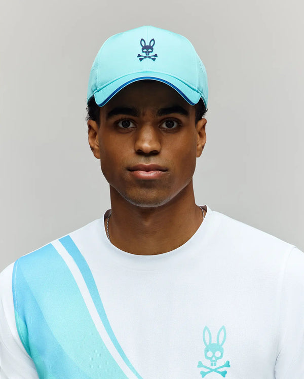 A person wears the MENS OFFICIAL US OPEN AVERY NET SPORT CAP by Psycho Bunny—a light blue, breathable hat with a bunny and crossbones logo—and a white shirt with blue stripes and matching bunny logo against a plain light gray background.