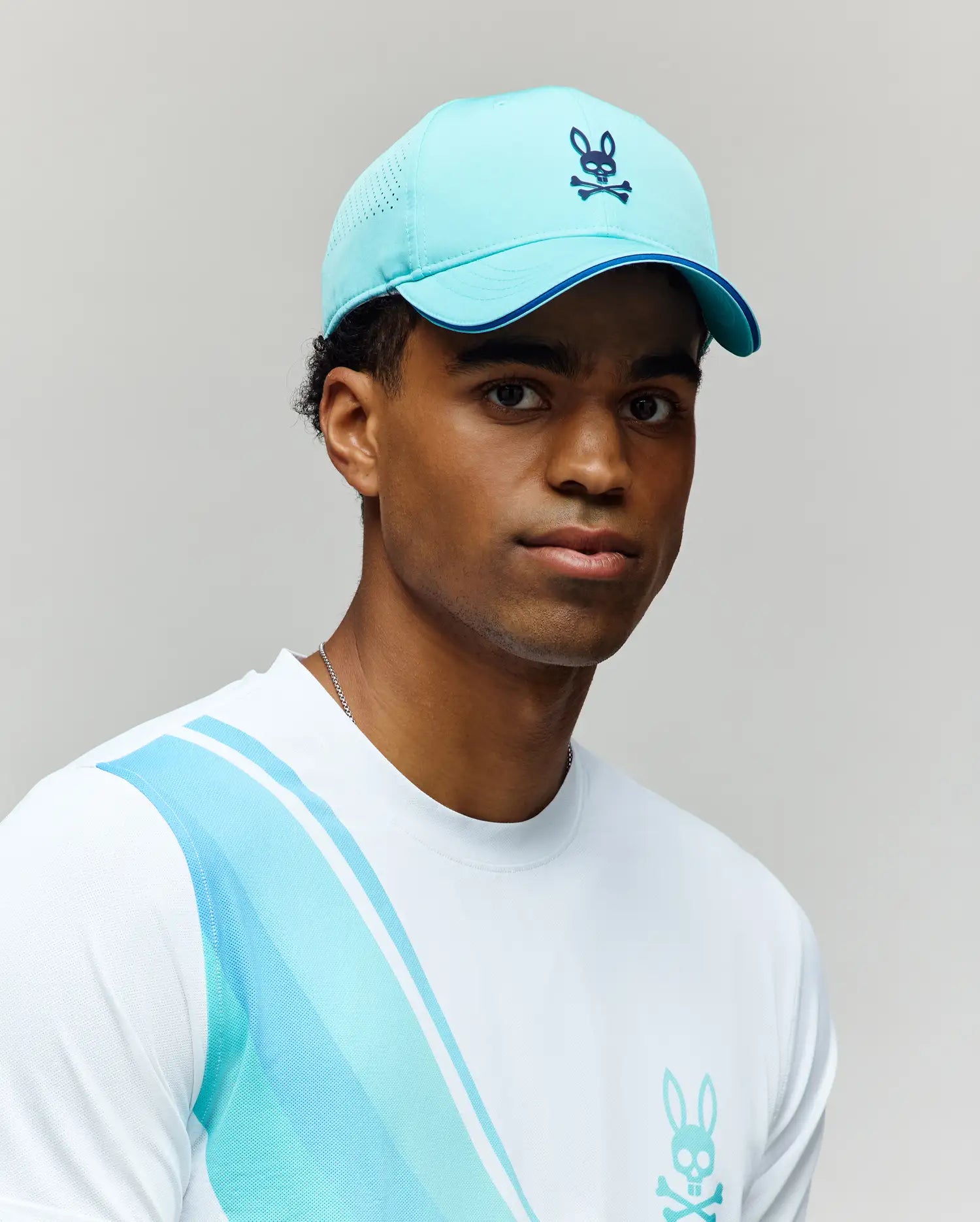 A young man stands against a light gray background wearing the Psycho Bunny MENS OFFICIAL US OPEN AVERY NET SPORT CAP in light blue, featuring a rabbit skull logo, along with a white shirt displaying a matching blue rabbit logo.