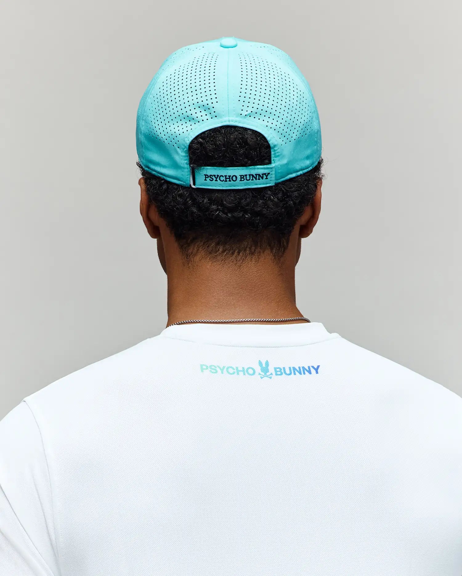 A person faces away, wearing the Psycho Bunny MENS OFFICIAL US OPEN AVERY NET SPORT CAP in turquoise, featuring the "PSYCHO BUNNY" logo above the strap and a white shirt with the blue and green "PSYCHO BUNNY" logo near the collar.