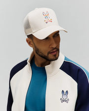 MENS RAPHAEL TRUCKER CAP, a cream trucker cap with a vibrant patterned Psycho Bunny skull logo, paired with a cream, navy, and teal color-blocked full-zip jacket featuring an embroidered bunny skull, over a teal crewneck t-shirt.