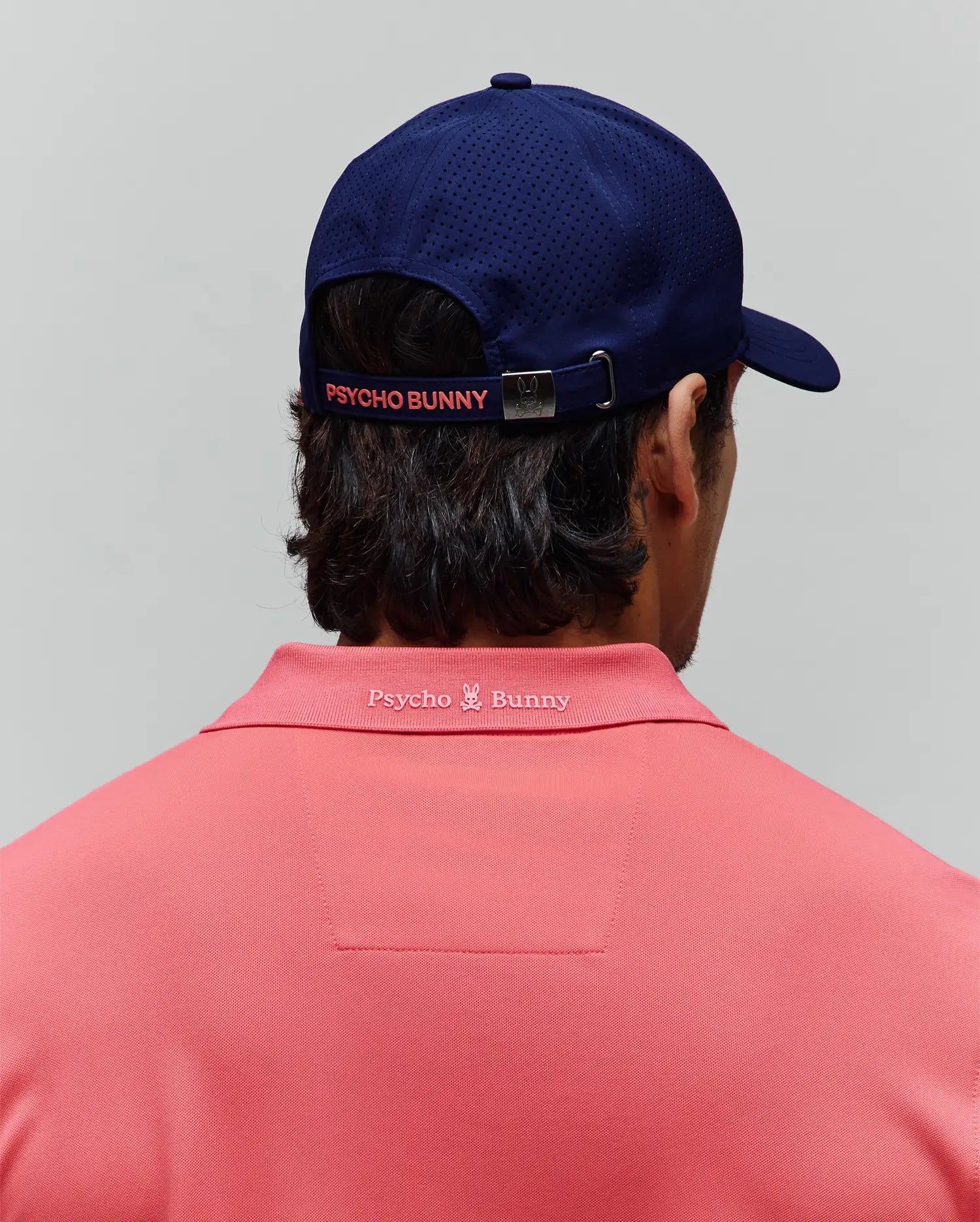 Coral pique polo with embroidered Psycho Bunny logo on back collar, paired with a navy perforated Mens Onyx Sport Cap featuring a coral Psycho Bunny strap and logo buckle.
