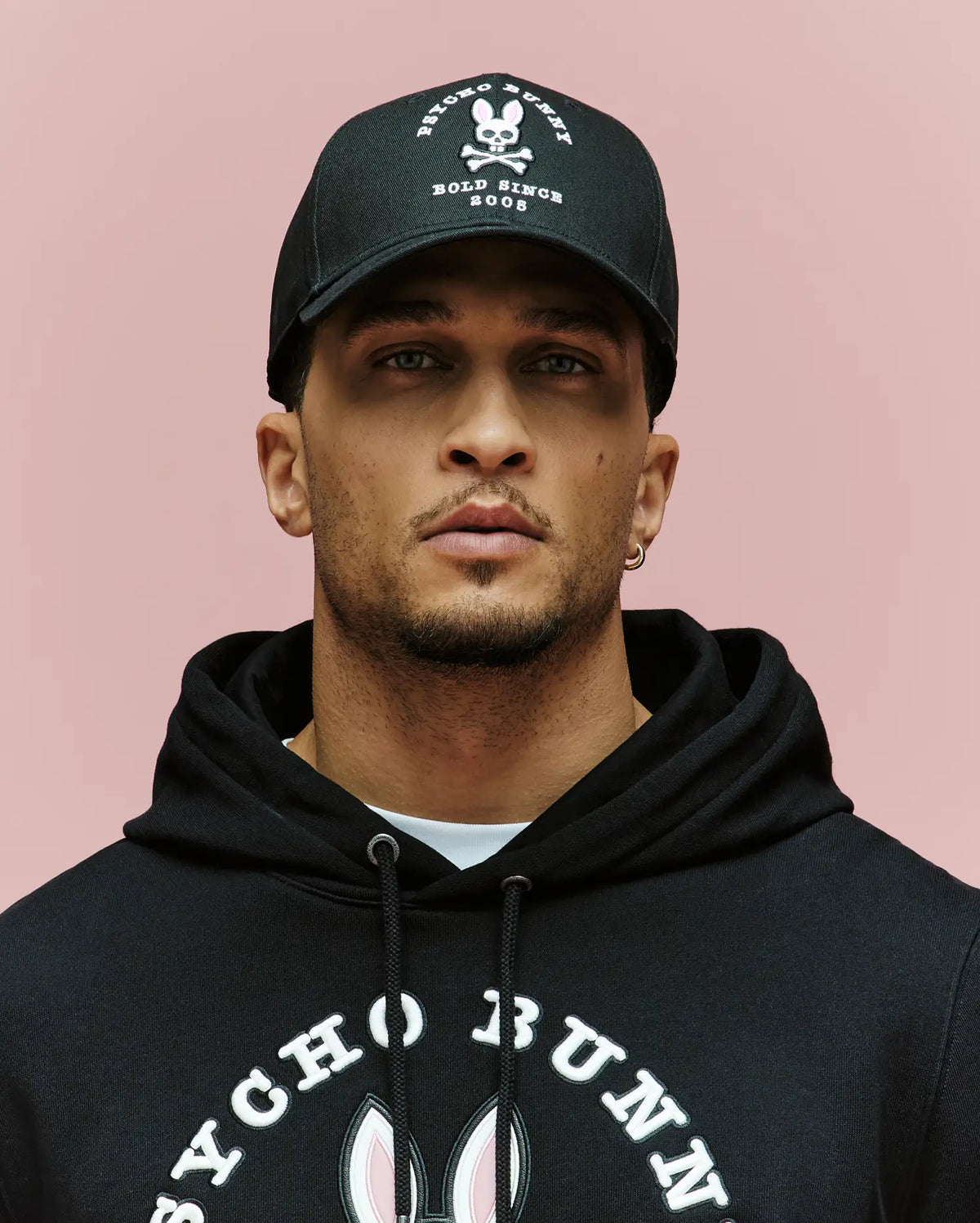 A man wearing the Psycho Bunny MENS ARCHIVE BASEBALL CAP - MO1500002, featuring an embroidered Bunny and skull and crossbones logo, and a black Psycho Bunny hoodie, poses against a plain pink background.