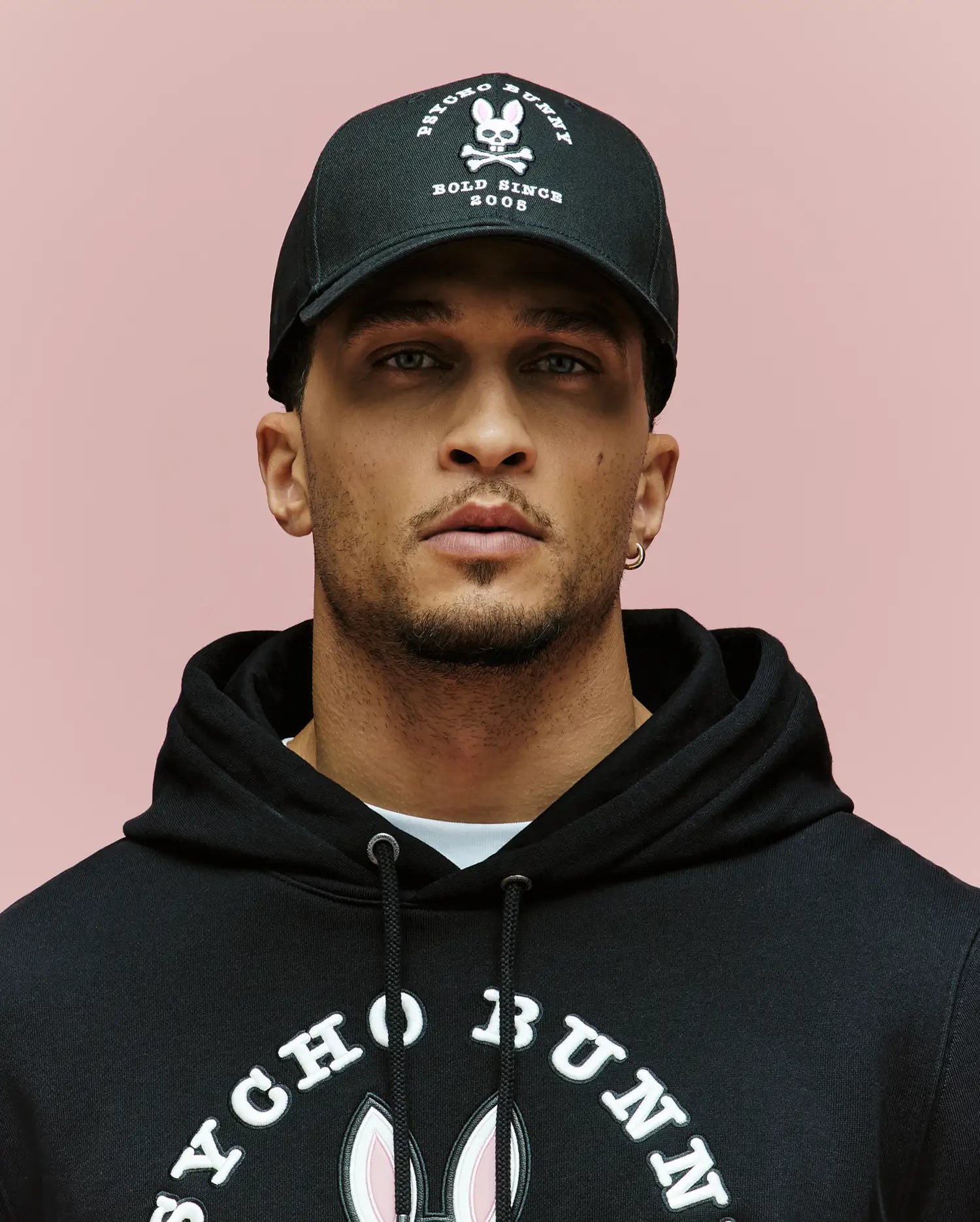 A man wearing the Psycho Bunny MENS ARCHIVE BASEBALL CAP - MO1500002, featuring an embroidered Bunny and skull and crossbones logo, and a black Psycho Bunny hoodie, poses against a plain pink background.