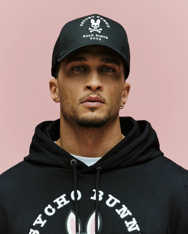 A man wearing the Psycho Bunny MENS ARCHIVE BASEBALL CAP - MO1500002, featuring an embroidered Bunny and skull and crossbones logo, and a black Psycho Bunny hoodie, poses against a plain pink background.