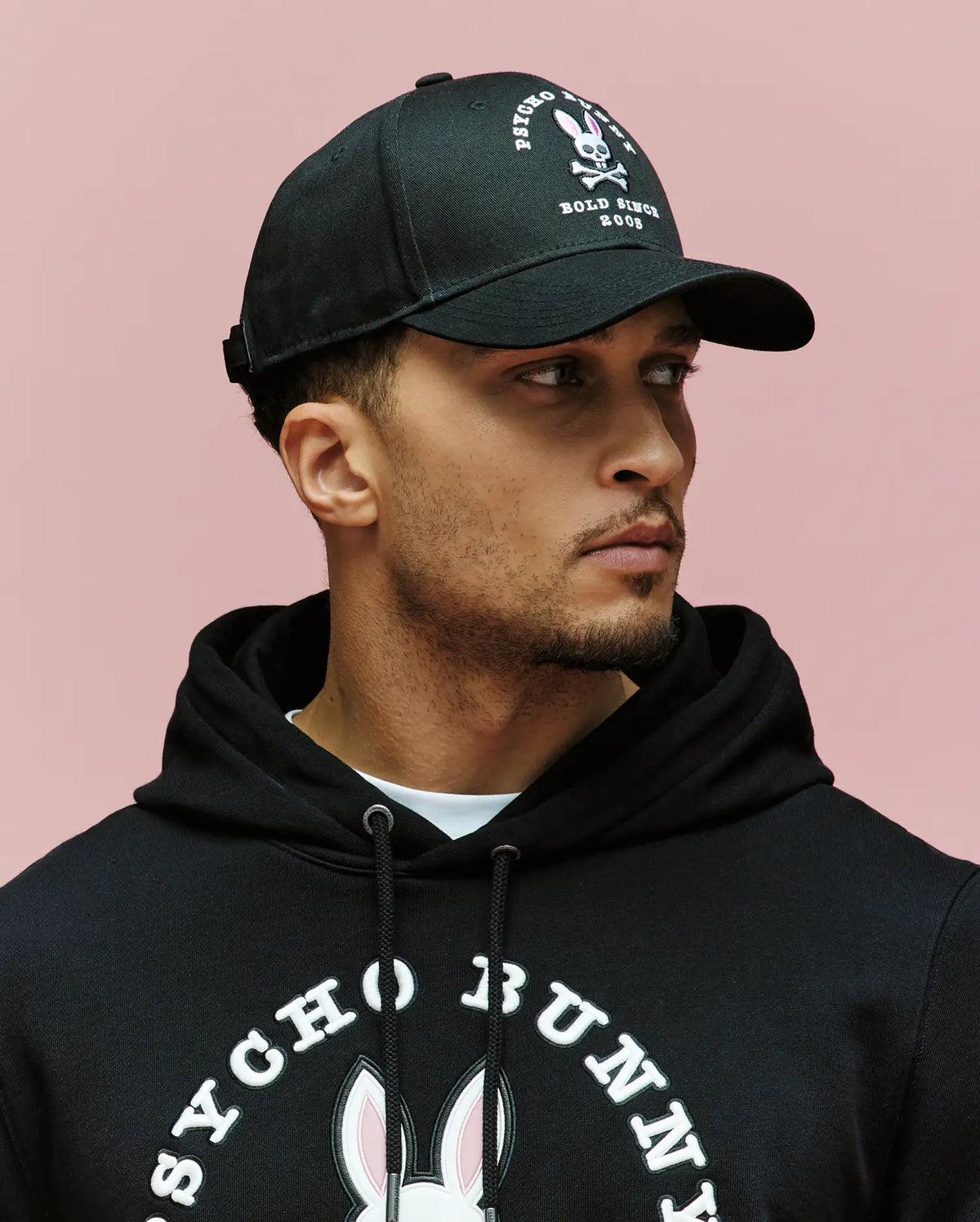 A man wearing a Psycho Bunny MENS ARCHIVE BASEBALL CAP - MO1500002 in black and a black hoodie with the embroidered Bunny logo and "Psycho Bunny" text stands against a pink background, looking to the side.