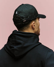 A person in a black hoodie and the Psycho Bunny MENS ARCHIVE BASEBALL CAP (MO1500002) with an adjustable strap is shown from behind against a light pink background. The cap displays a small embroidered Bunny logo on the back.